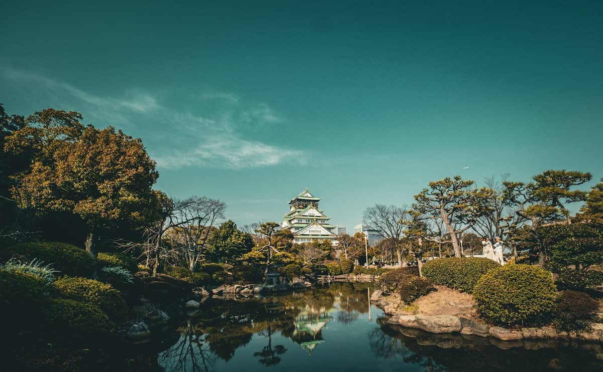 Osaka Castle reflected in a calm pond, surrounded by manicured trees and lush greenery under a clear blue sky.