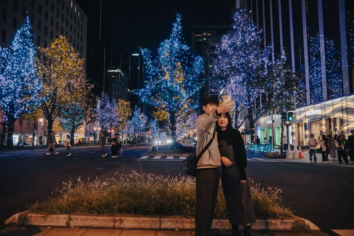 A couple taking a selfie at night surrounded by blue and yellow illuminated trees in a city street in Osaka.