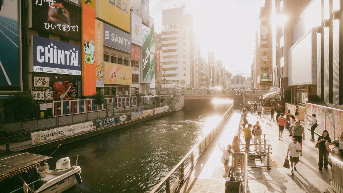 Sunlit view of Osaka's Dotonbori canal with people walking along the waterfront and colorful building signs lining the waterway.