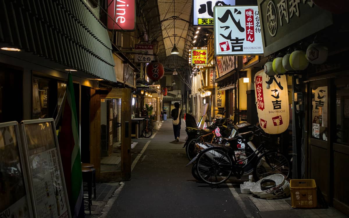 Atmospheric night view of a nostalgic Osaka alleyway lined with izakayas, paper lanterns, and bicycles, showcasing a hidden side street that locals enjoy.