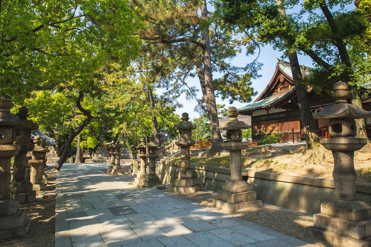 Stone lanterns line a shaded path leading to a traditional wooden shrine in a quiet Osaka temple setting.