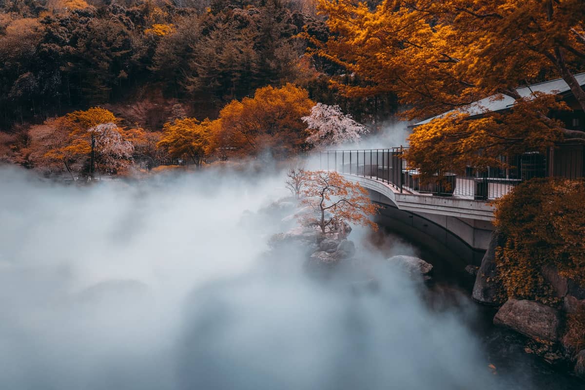 Autumn foliage in vivid orange and yellow around a bridge with mist over a river, forested hills in the background.