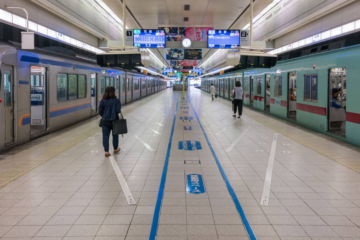 Bright indoor train platform in Osaka with two trains, blue directional signs, and commuters walking along the tiled floor.