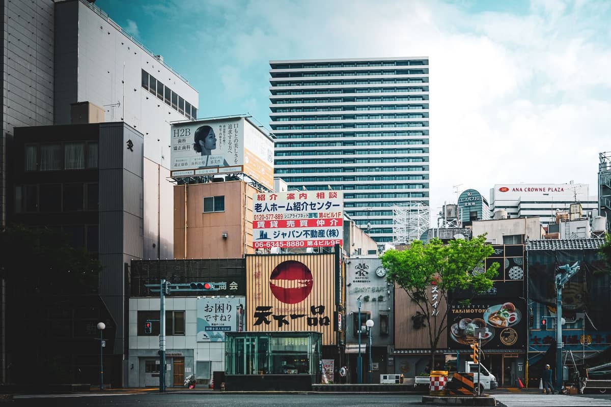 Cityscape of central Osaka with modern buildings, commercial signs, and light clouds in a bright sky, showing typical urban autumn weather.