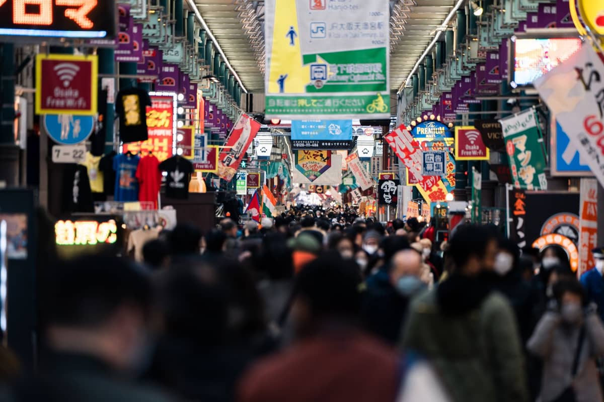 A crowded, colorful indoor shopping street in Osaka, packed with people under banners and store signs indicating high tourist activity.