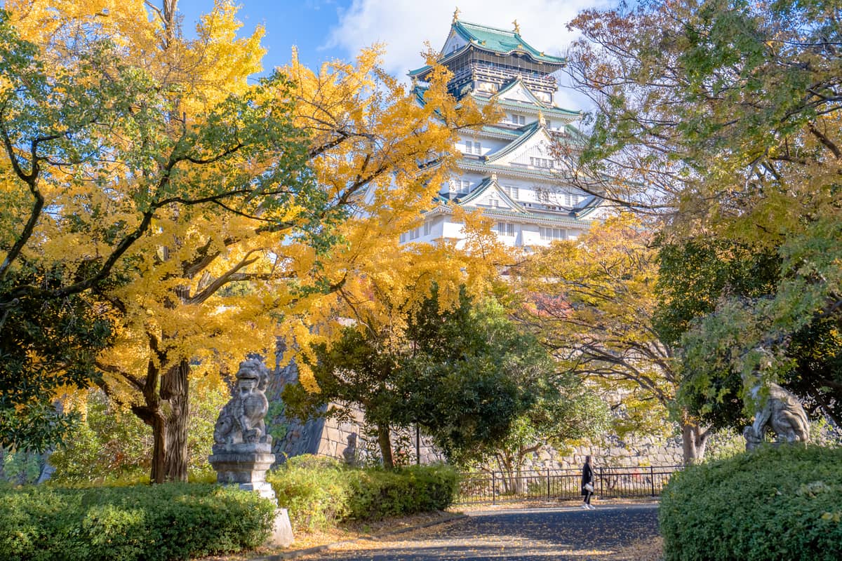 Osaka Castle framed by vibrant autumn foliage and stone statues, with a clear blue sky on a sunny fall day.