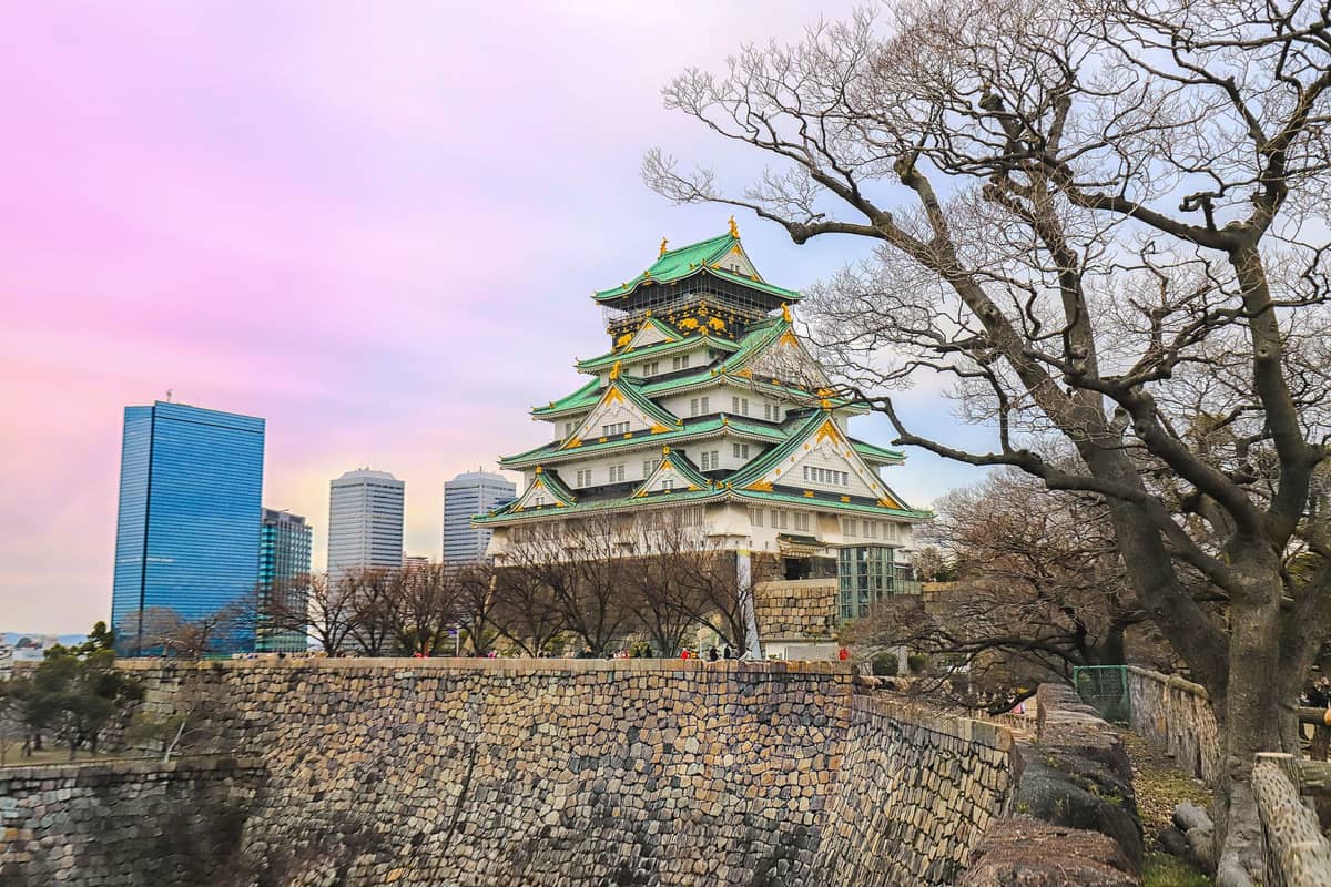 Osaka Castle with bare winter trees in front, city skyscrapers in the background, and a soft pink sky during winter.