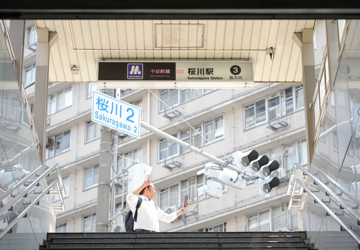 A person takes a selfie at the entrance of Sakuragawa Station in Osaka, showing street signs and city buildings.