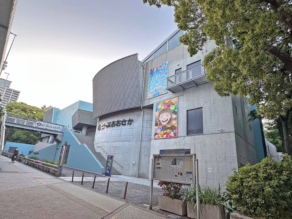 Exterior view of Osaka International Peace Center (Peace Osaka) with blue and gray modern architecture, Japanese signage, colorful murals, and trees in front.