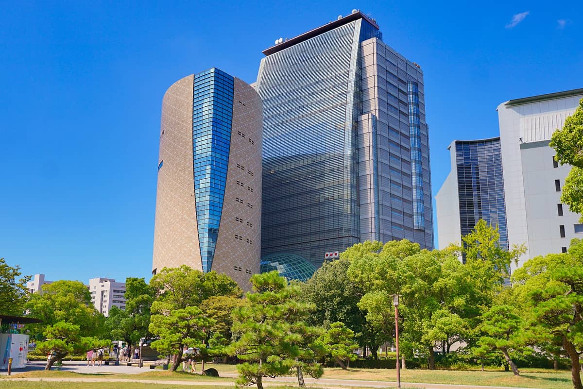 The modern, angular exterior of the Osaka Museum of History adjacent to a glass office tower, with trees and a clear blue sky in front.