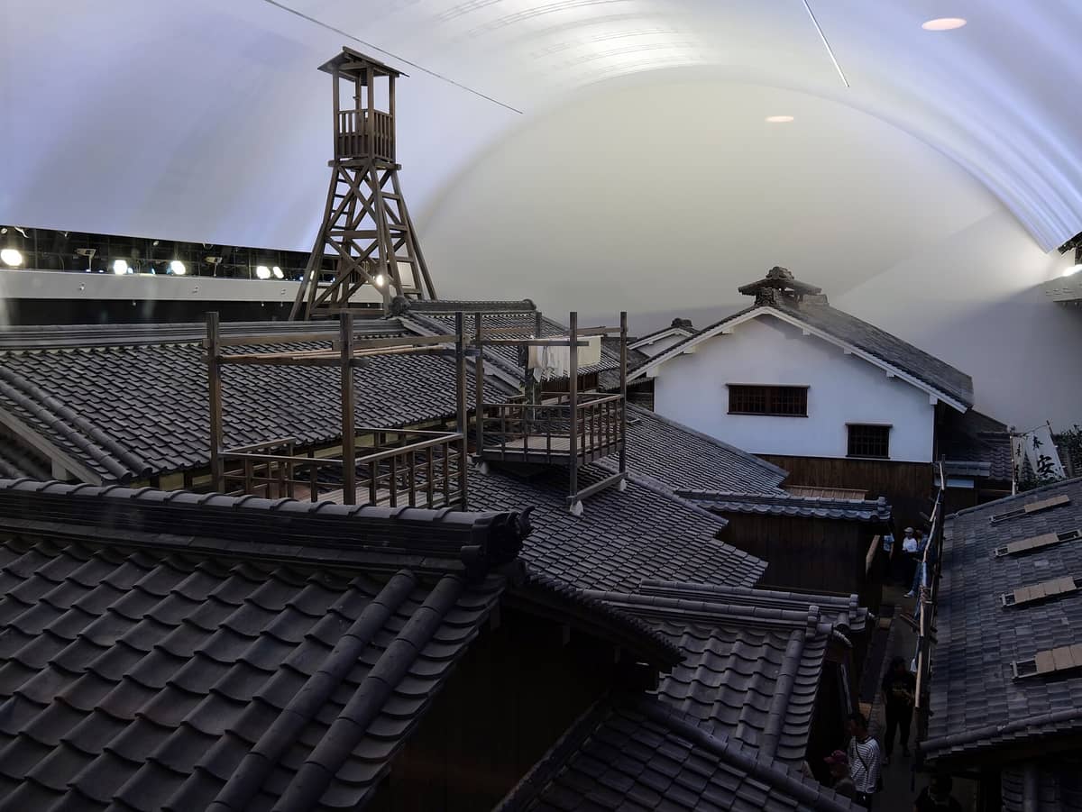 Edo-period style merchant homes and a wooden fire lookout tower reconstructed inside the Osaka Museum of Housing and Living, viewed from above under an arched modern ceiling.