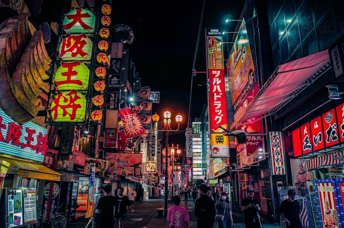 Bustling Osaka nightlife street filled with neon lights, colorful shopfronts, and people walking under bright signs in Japanese at night.