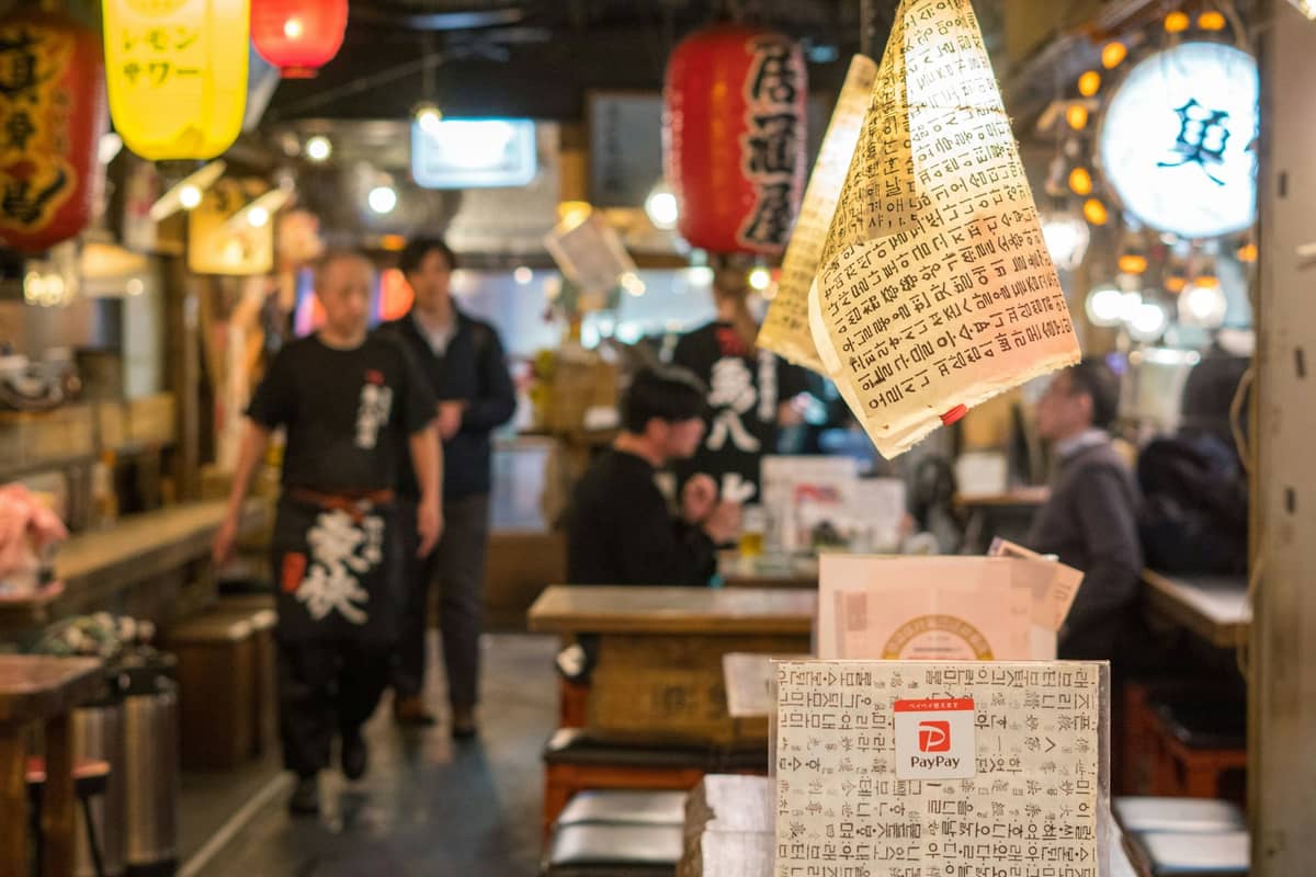 Interior of a lively izakaya with hanging lanterns, wooden tables, customers dining and staff moving through the warmly lit space.