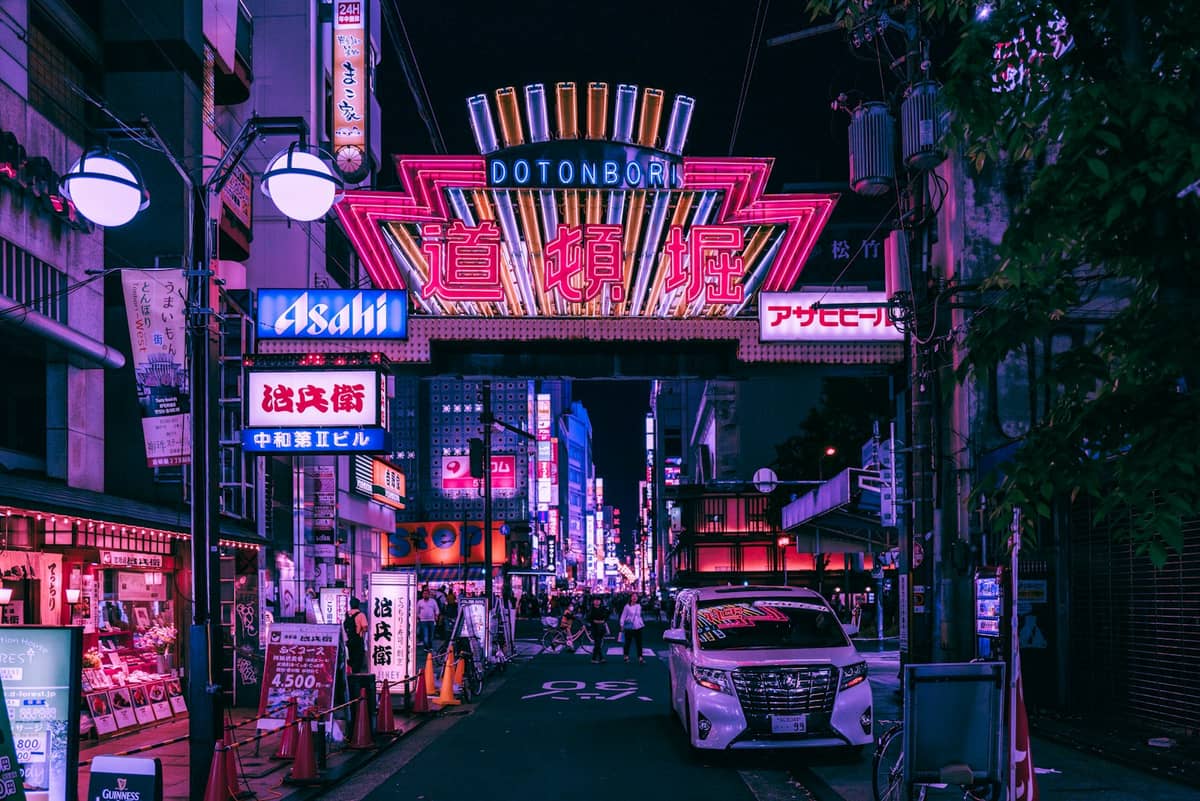 Night view of Osaka's Dotonbori street with its iconic neon-lit entrance sign, vibrant nightlife, shops, and taxis under glowing signage.