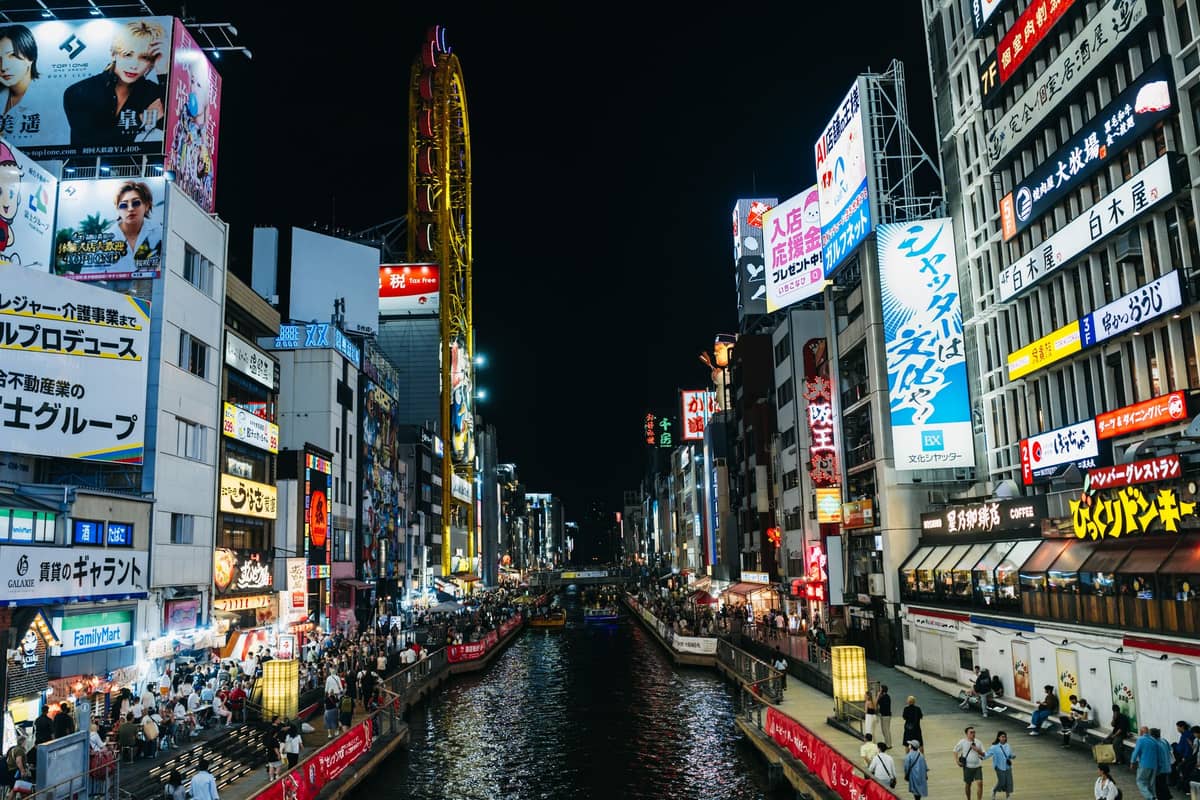 Osaka’s Dotonbori canal at night, lined with brightly lit buildings, colorful billboards, and bustling crowds along both embankments.