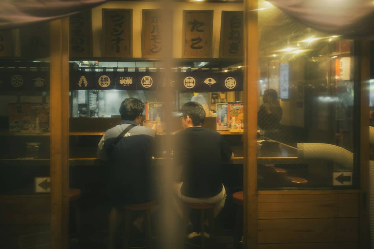 Two people sitting at a counter inside a cozy Japanese restaurant at night, seen through a window, with warm lighting and food signs above.