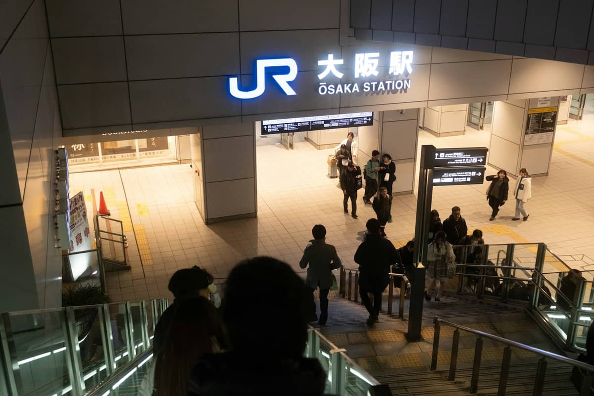 People descending a stairway toward the JR Osaka Station entrance, with a bright sign reading Osaka Station at night.