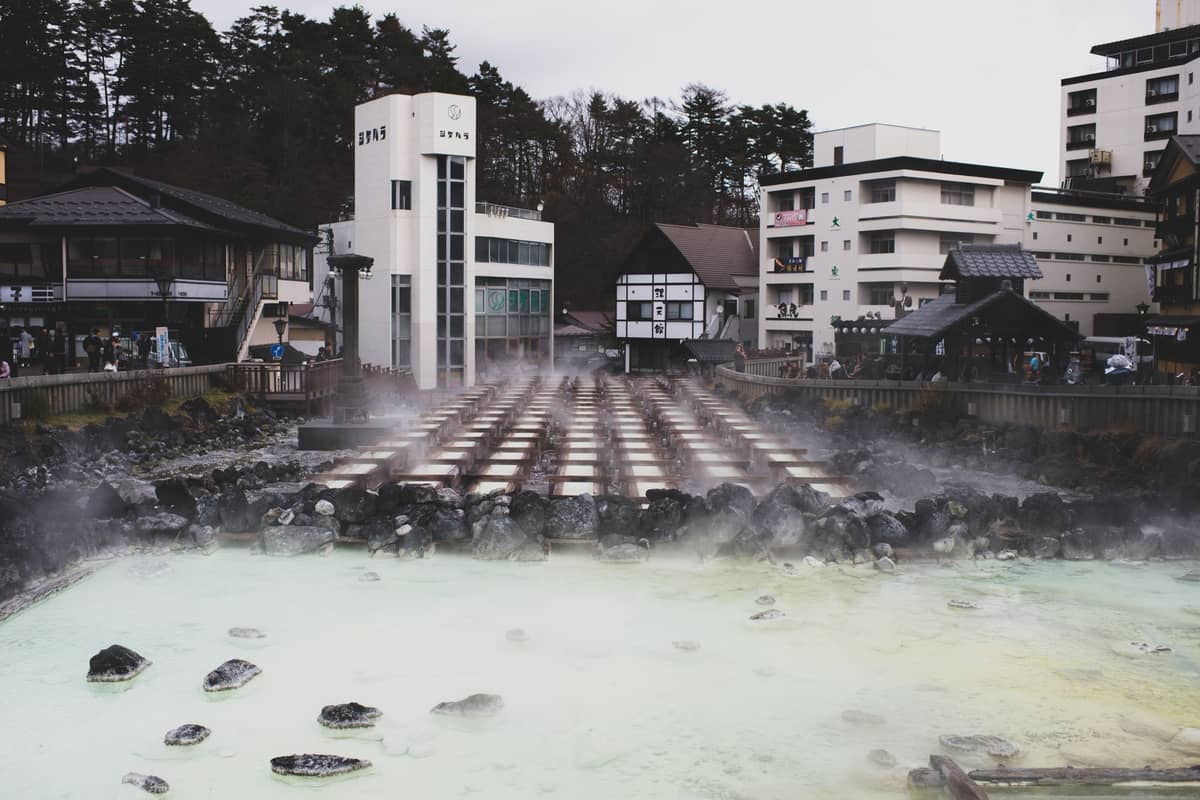 Steam rising from a milky-colored outdoor onsen pool surrounded by urban buildings, likely in a Japanese hot spring town.