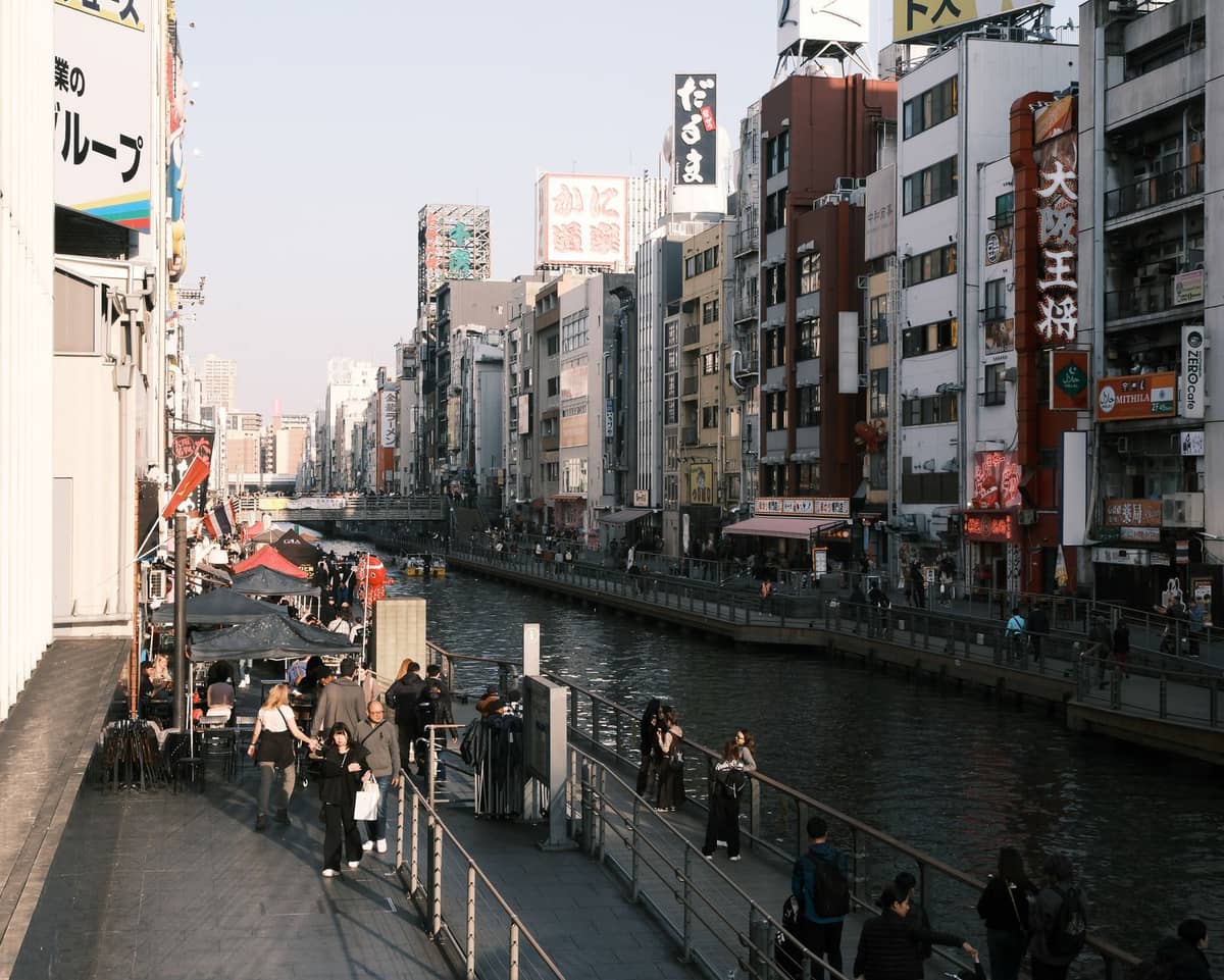 Daytime view of Dotonbori canal in Osaka with busy pedestrian walkways, colorful buildings, and vibrant signage lining both sides of the water.