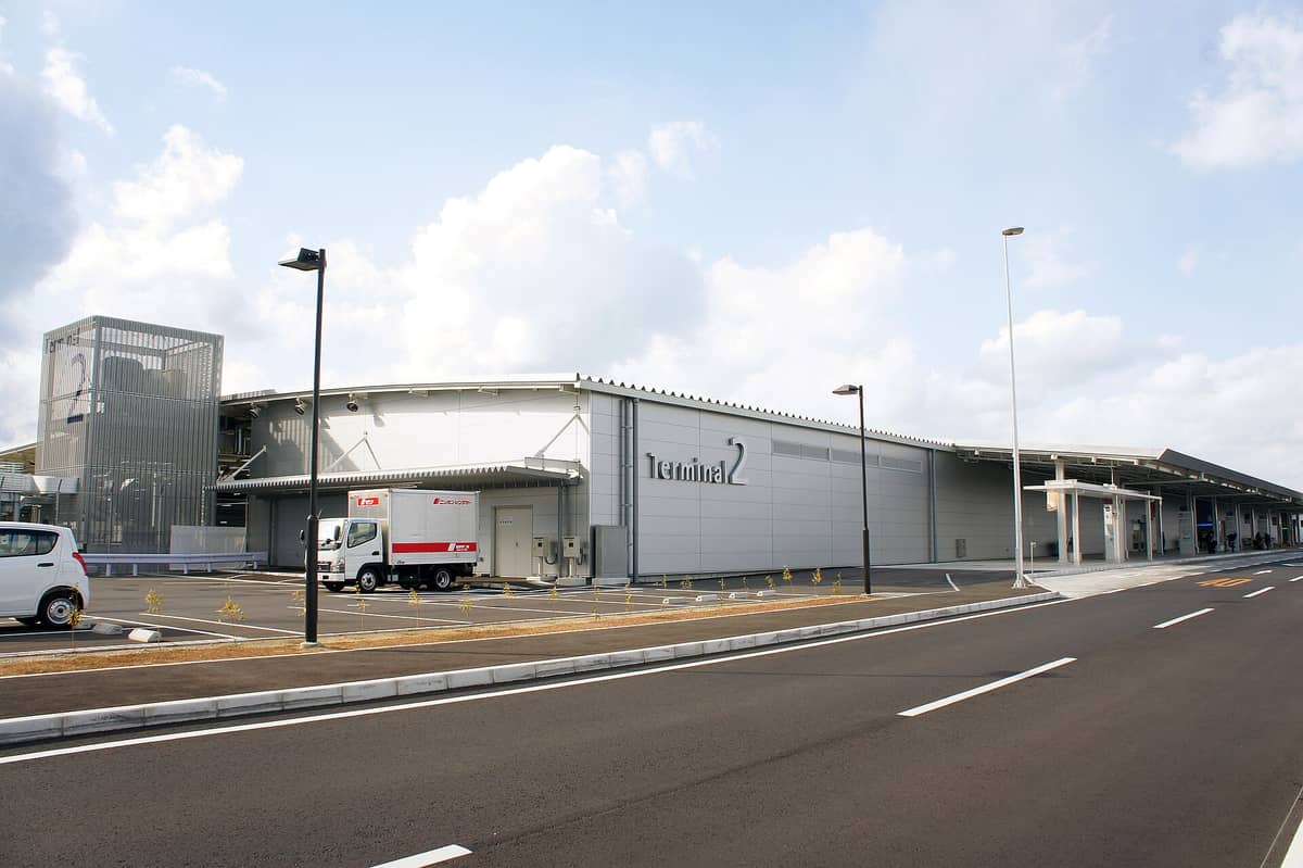 Exterior view of Kansai International Airport Terminal 2 in Osaka with parking and signage visible under a partly cloudy sky.