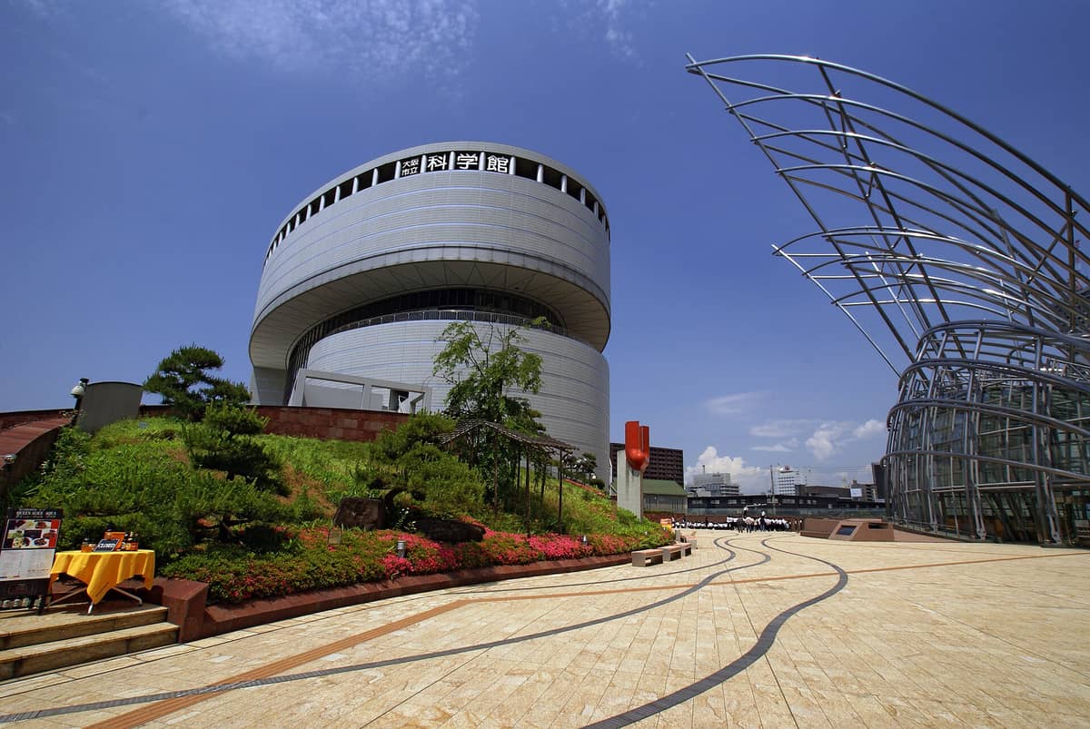 Exterior view of the Osaka Science Museum, featuring its distinctive round planetarium building and modern architectural elements under a blue sky.