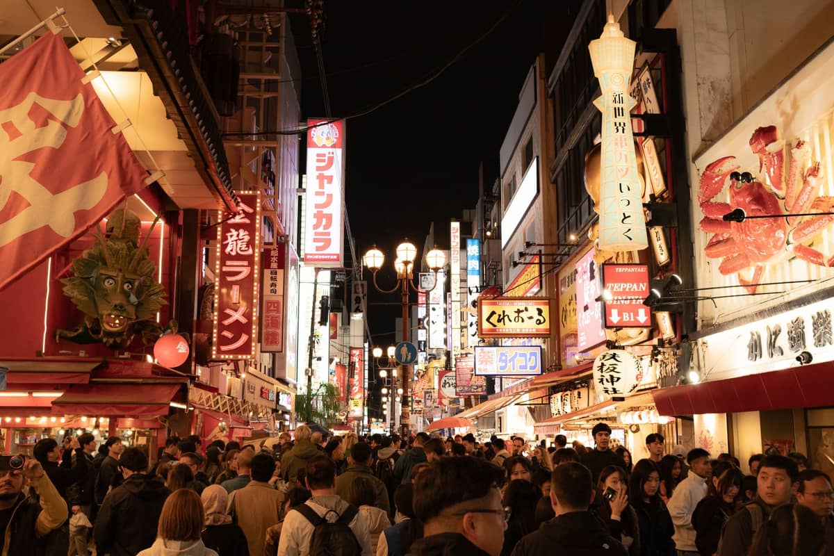 Bustling night scene of Dotonbori in Osaka, crowded with shoppers and bright neon shop signs covering buildings on both sides.