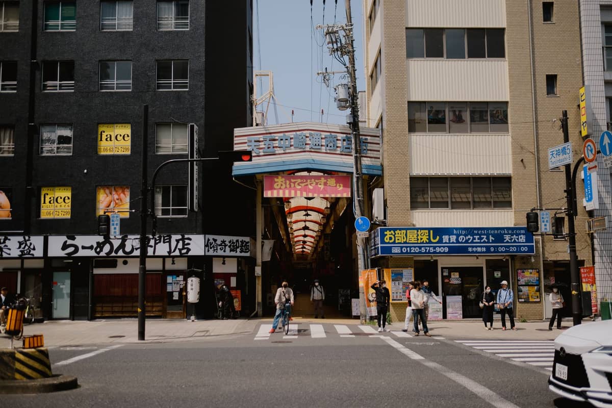 Street view of an entrance to a Japanese shopping arcade with people crossing and shop signs in Japanese, likely in Osaka.