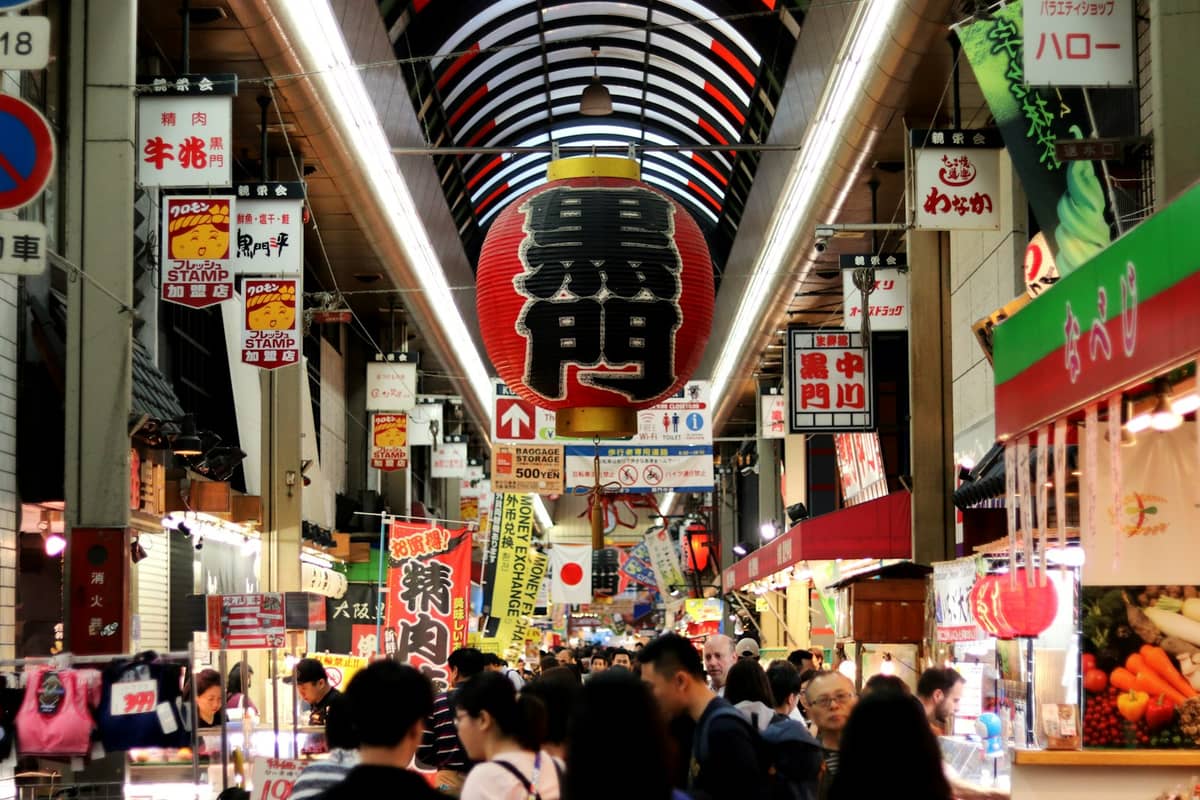 Crowded indoor market arcade with a large red lantern, Japanese signs, and shoppers exploring food stalls at Kuromon Ichiba Market, Osaka.