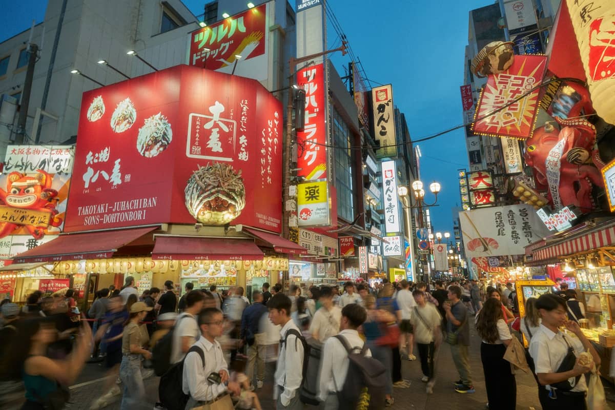 Crowded street in Osaka’s famous Dotonbori district at dusk, with bright restaurant signs and street food stalls lining both sides.