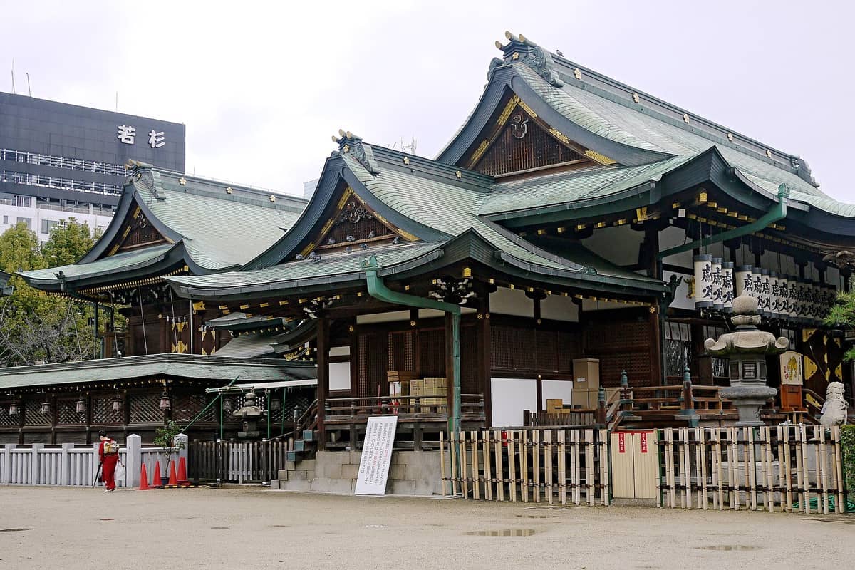 Traditional wooden buildings of Osaka Tenmangu Shrine with green-tiled roofs and a stone lantern, set against a cloudy sky in Osaka.