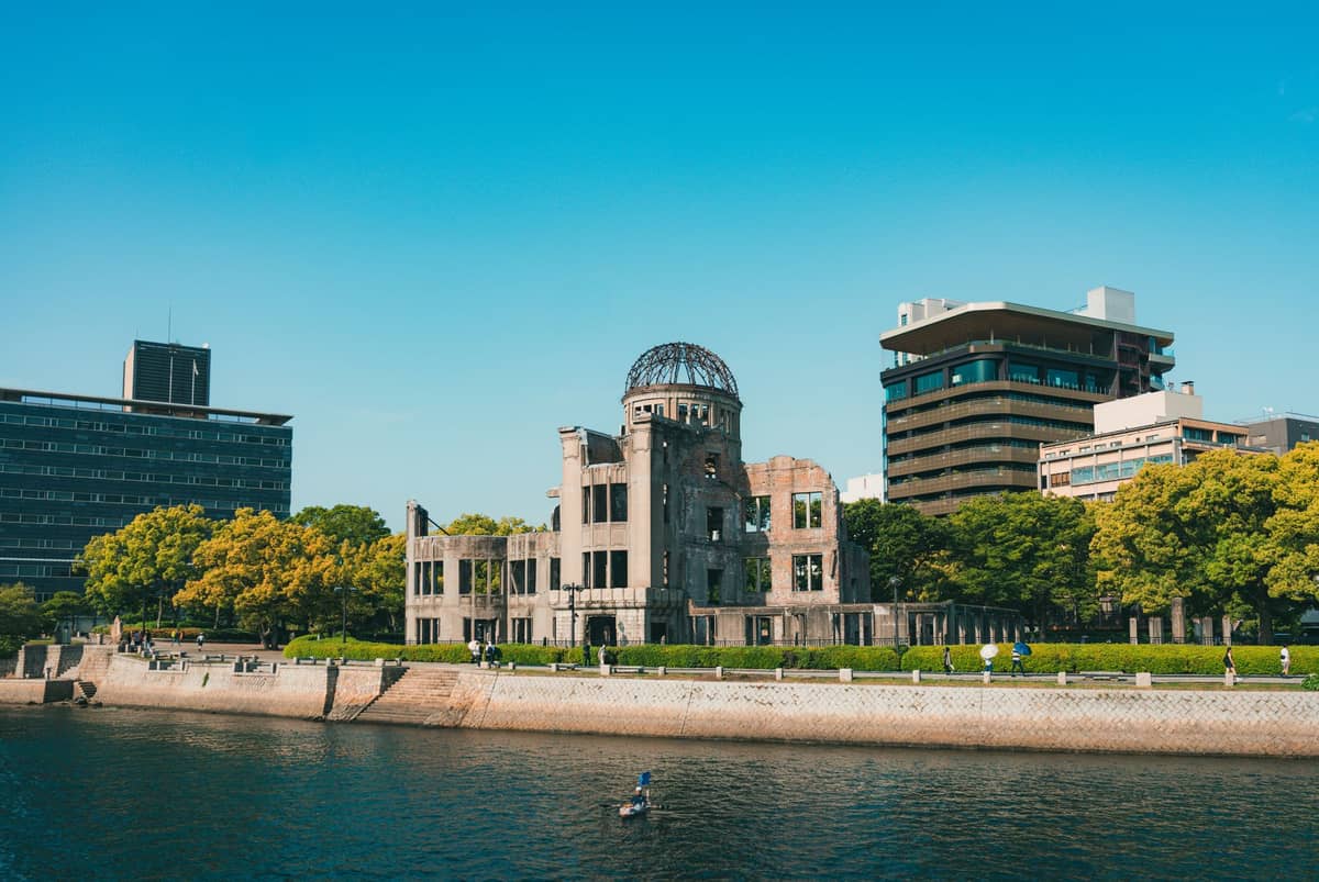 The Hiroshima Peace Memorial (Atomic Bomb Dome) beside the Motoyasu River, surrounded by greenery and city buildings under a clear blue sky.