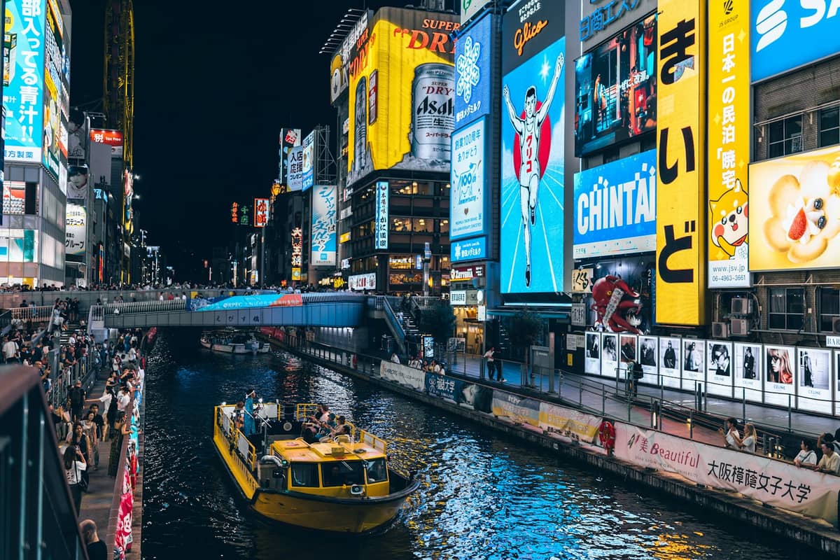 Night view of Osaka's Dotonbori canal with bright neon lights, colorful billboards, busy walkways, and a yellow tour boat on the water.