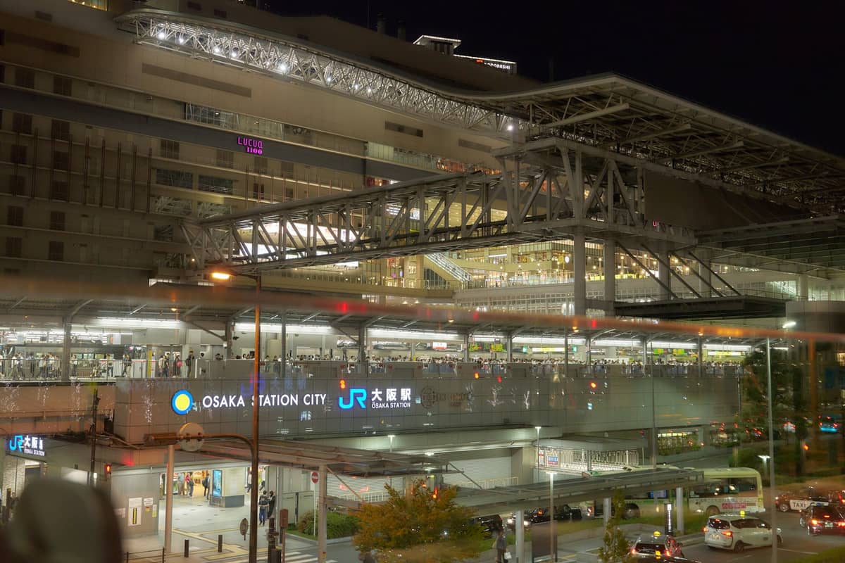 Night view of Osaka Station City with illuminated JR signs, pedestrian skybridge, and surrounding urban buildings.