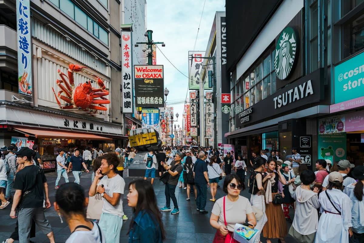 Busy Osaka street scene in Dotonbori with crowds of people, colorful storefronts, and famous crab restaurant sign.