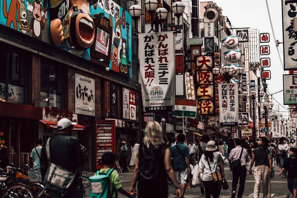 Crowded Dotonbori street scene in Osaka with people walking and colorful neon shop signs and advertisements lining both sides.