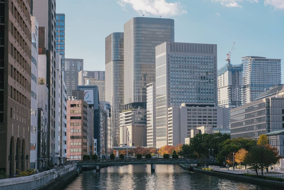 Modern buildings line both sides of a canal in central Osaka with a bridge in the distance and reflections in the water.