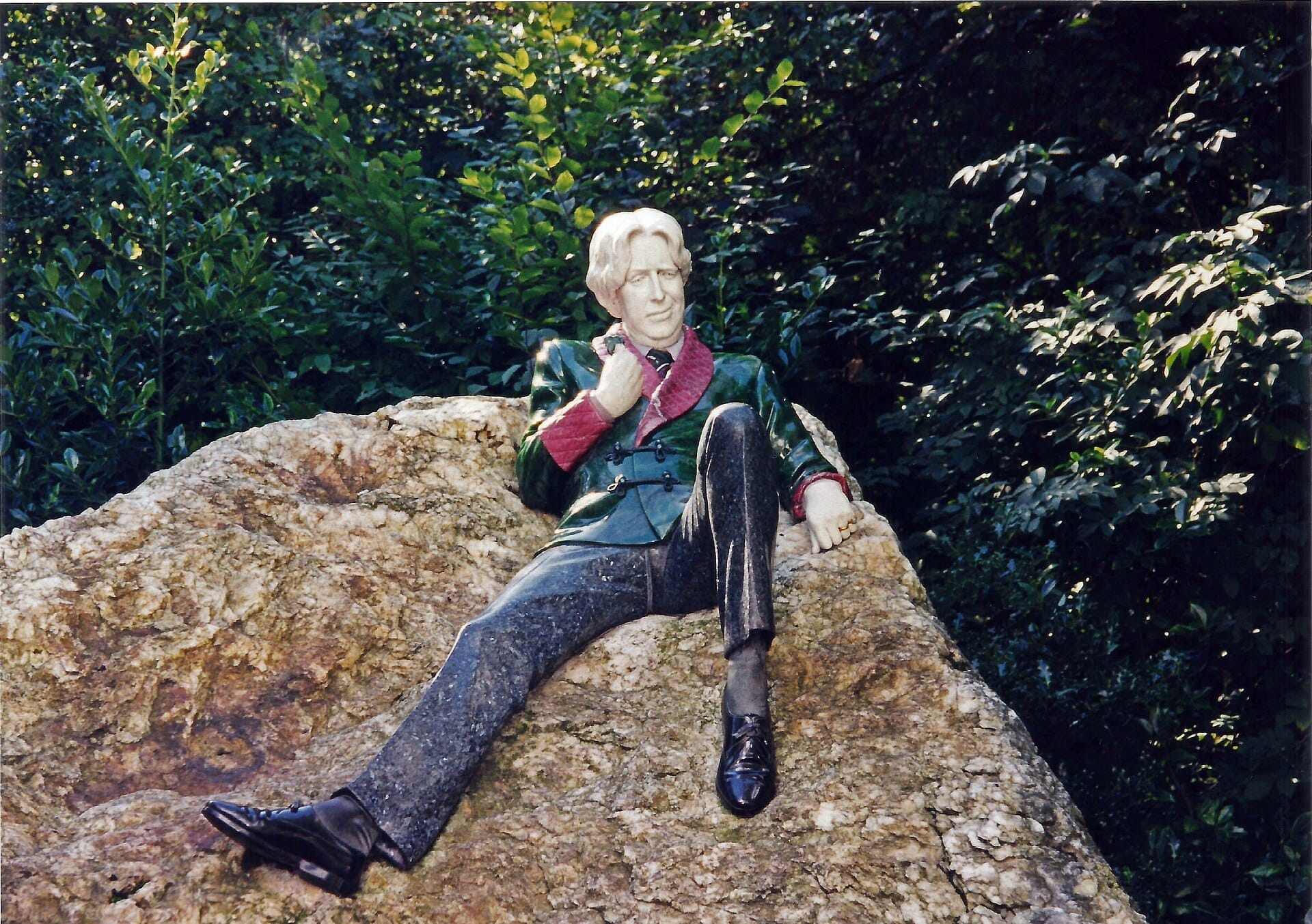 Scultura commemorativa di Oscar Wilde sdraiata su un grande masso di quarzo a Merrion Square, circondata da una lussureggiante vegetazione verde alla luce naturale del giorno.
