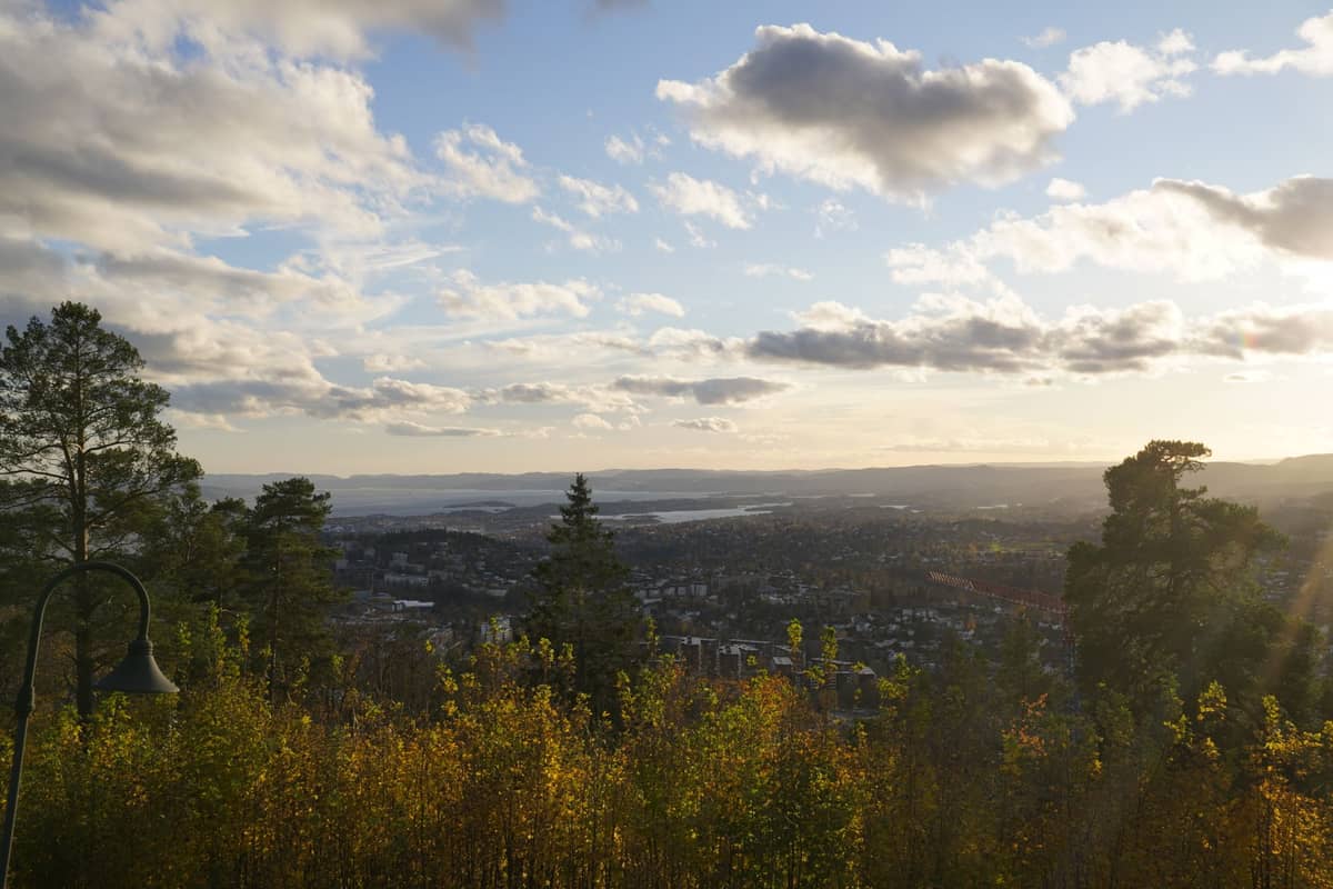 วิวกว้างจากไหล่เขาที่มีต้นไม้ปกคลุมมองลงไปยัง Oslo และ Oslofjord พร้อมต้นไม้ ทิวทัศน์เมือง เมฆกระจาย และแสงอาทิตย์ยามเย็น