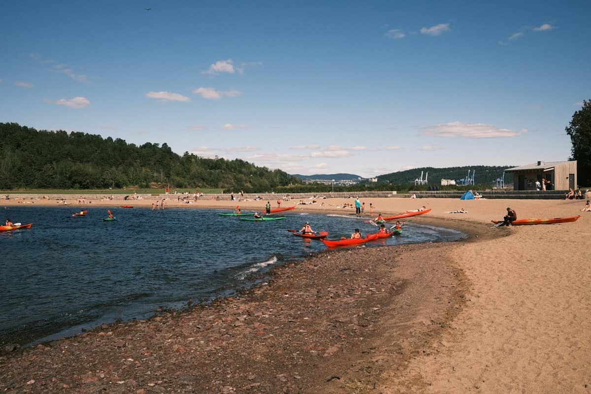 ชายหาดทรายริม Oslofjord พร้อมผู้คนที่อาบแดดและพายเรือคายัค ล้อมรอบด้วยป่าและอาคารในเมืองเป็นฉากหลัง