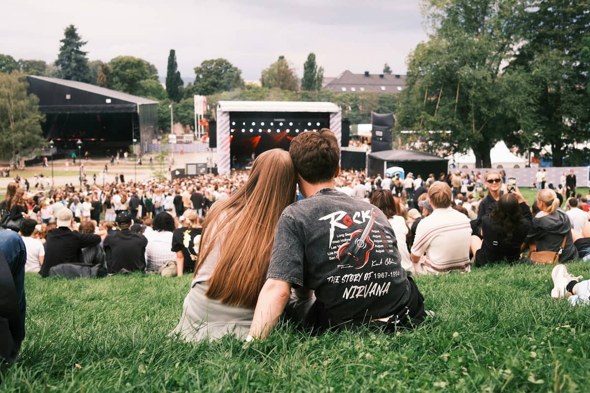 คู่รักหนุ่มสาวนั่งบนสนามหญ้าในงานเทศกาลดนตรีกลางแจ้งขนาดใหญ่ที่ Oslo โดยมีฝูงชน เวที และต้นไม้เป็นฉากหลัง