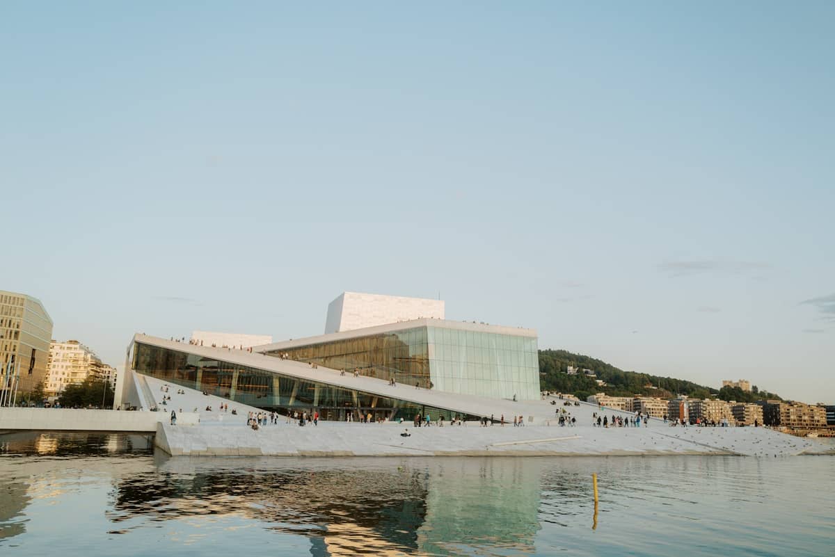 โรงอุปรากร Oslo พร้อมหลังคาหินอ่อนลาดเอียงที่เดินได้และผู้คน มองเห็นจากริมน้ำในวันที่อากาศสงบ