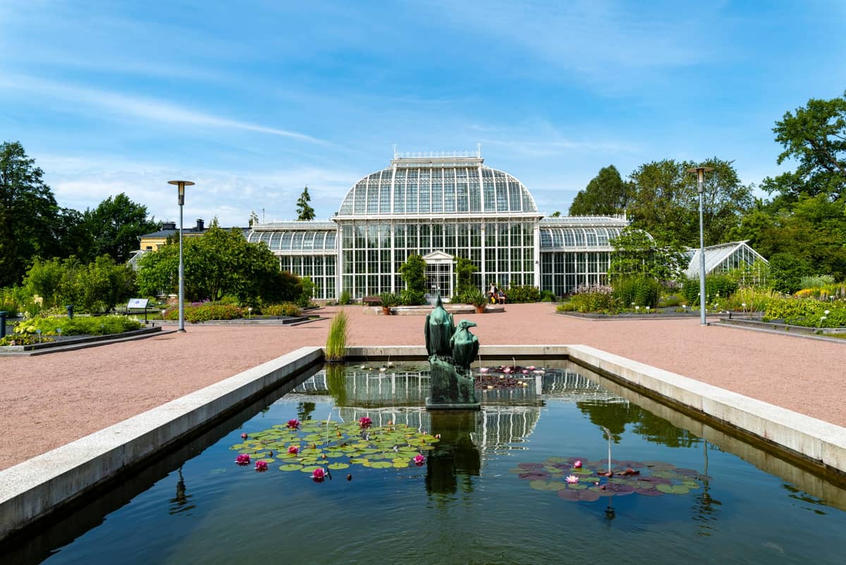เรือนกระจกในสวนพฤกษศาสตร์ Oslo สระน้ำสะท้อนแสงพร้อมรูปปั้นด้านหน้า ล้อมรอบด้วยสวนดอกไม้บานสะพรั่งในวันที่แสงแดดสดใส