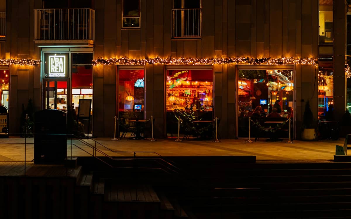 หน้าบาร์ Døgnvill Bar & Burger ในออสโลยามค่ำ แสงไฟอบอุ่น แขกในร้านและไฟนอกอาคาร ลงตัวกับบรรยากาศกลางเมืองที่เต็มไปด้วยชีวิตยามค่ำคืน