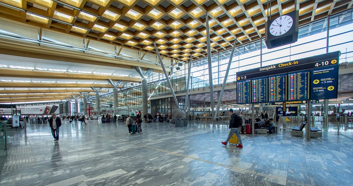 ภายในกว้างขวางของสนามบิน Oslo Gardermoen พร้อมป้ายบอกเที่ยวบินขาออกขนาดใหญ่ สถาปัตยกรรมไม้และกระจกสมัยใหม่ ผู้โดยสาร และสัมภาระ