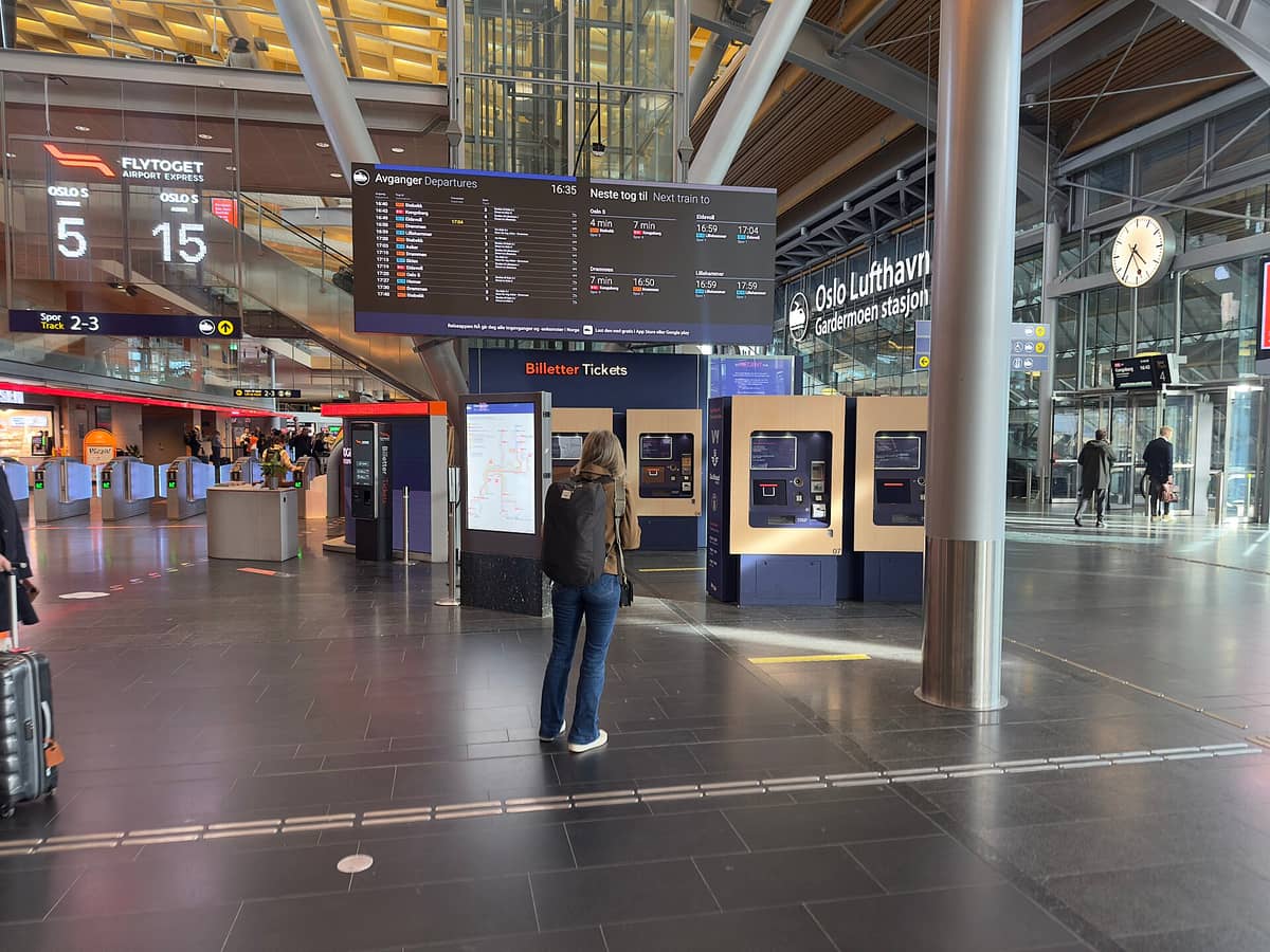 ภายในสถานีรถไฟสนามบินออสโลพร้อมเครื่องจำหน่ายตั๋ว จอแสดงตารางออกเดินทาง และป้ายบอกทางของ Flytoget Airport Express และ Oslo Lufthavn Gardermoen