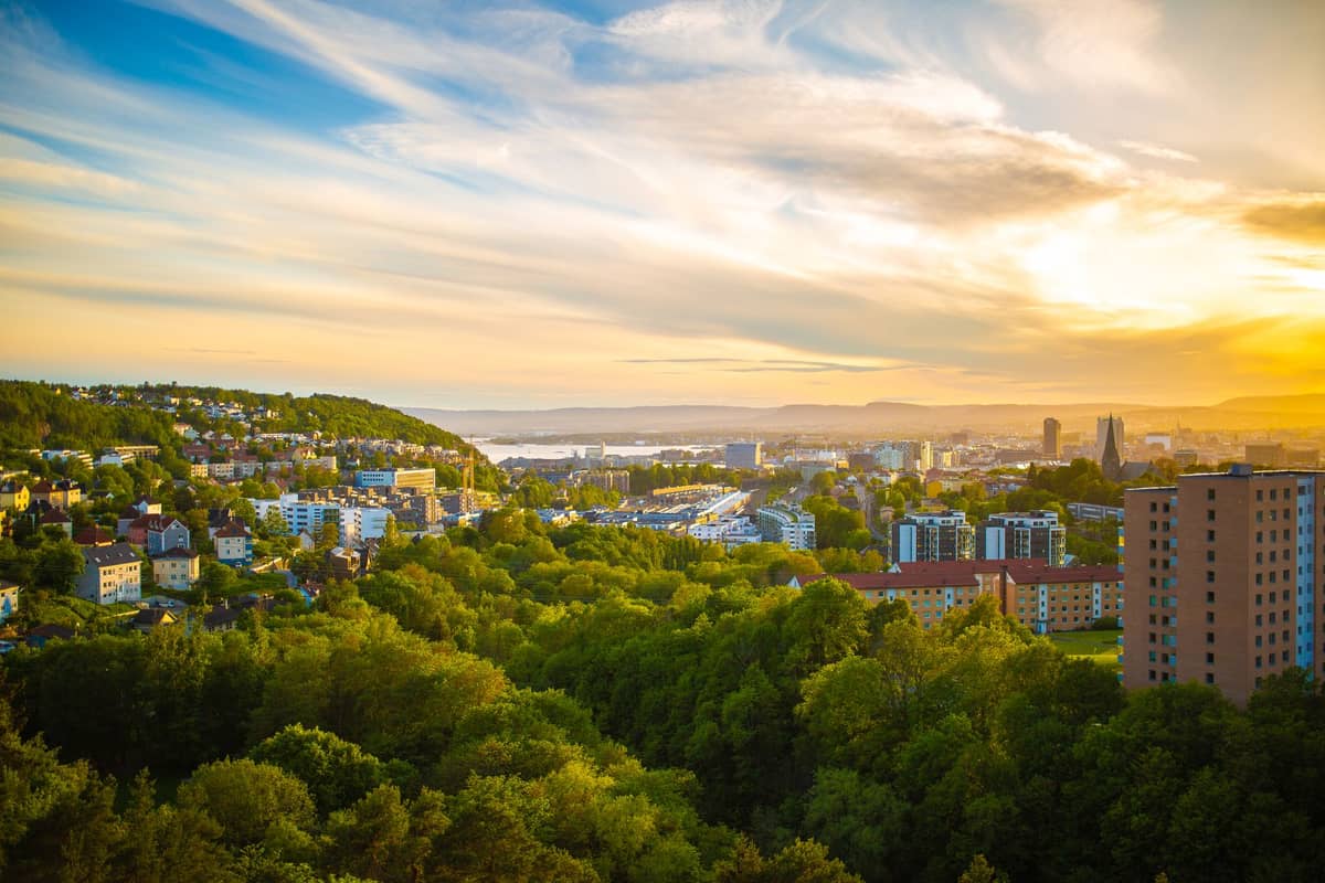 วิวกว้างของ Oslo โดยมีสวนสาธารณะสีเขียวชอุ่มอยู่เบื้องหน้า อาคารของเมืองและเนินเขาอยู่เบื้องหลังในยามพระอาทิตย์ตก
