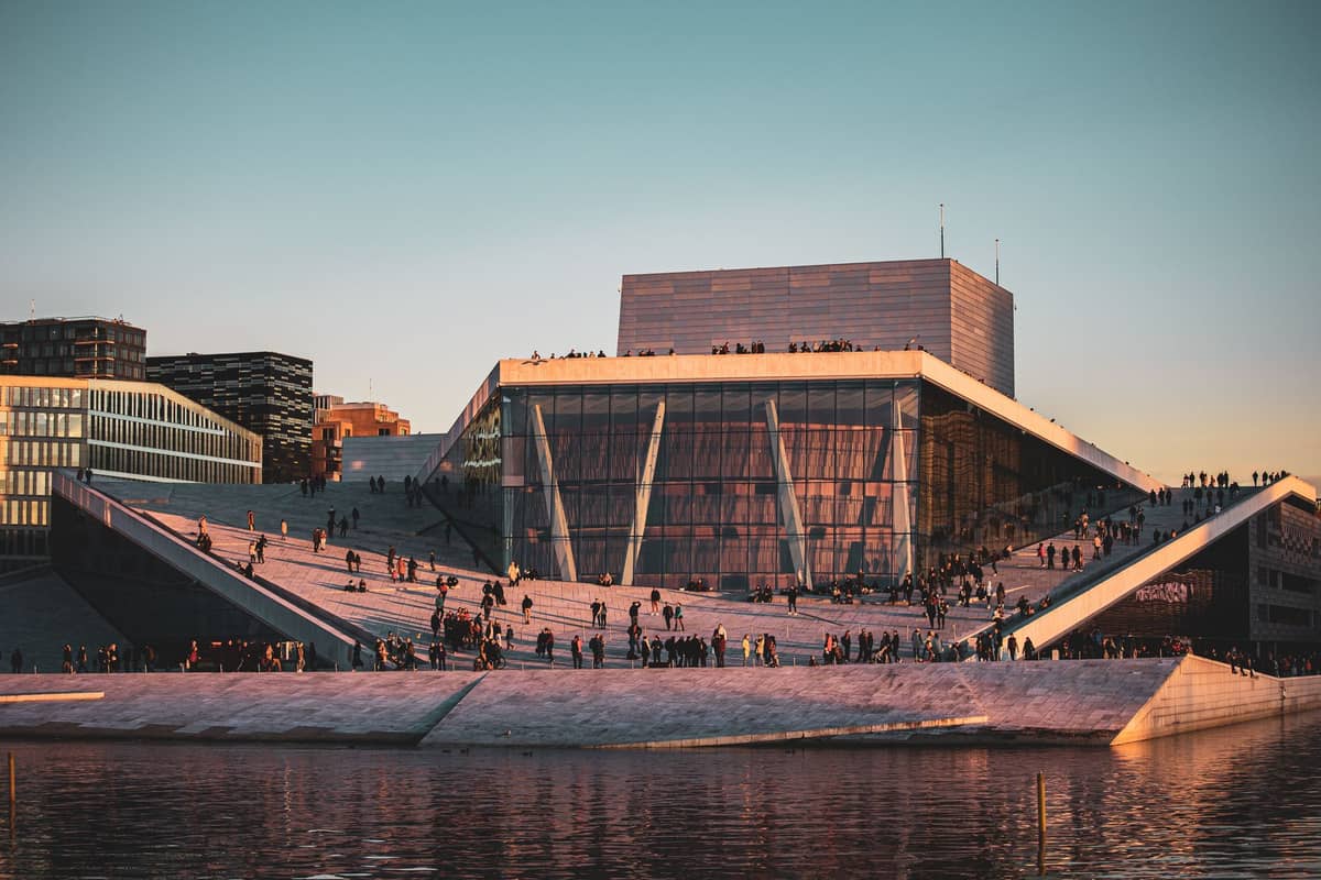 Oslo Opera House ยามพระอาทิตย์ตกดินพร้อมผู้คนที่เดินบนหลังคาเอียงและอาคารริมน้ำในพื้นหลัง