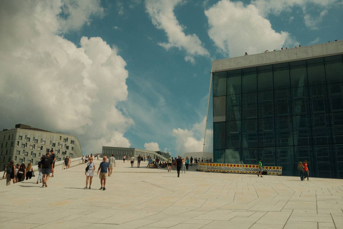 ผู้คนเดินและรวมตัวกันบนหลังคาหินอ่อนลาดเอียงของ Oslo Opera House ใต้ท้องฟ้าสีฟ้าที่มีเมฆบางส่วน โดดเด่นด้วยจุดชมวิวทางสถาปัตยกรรม