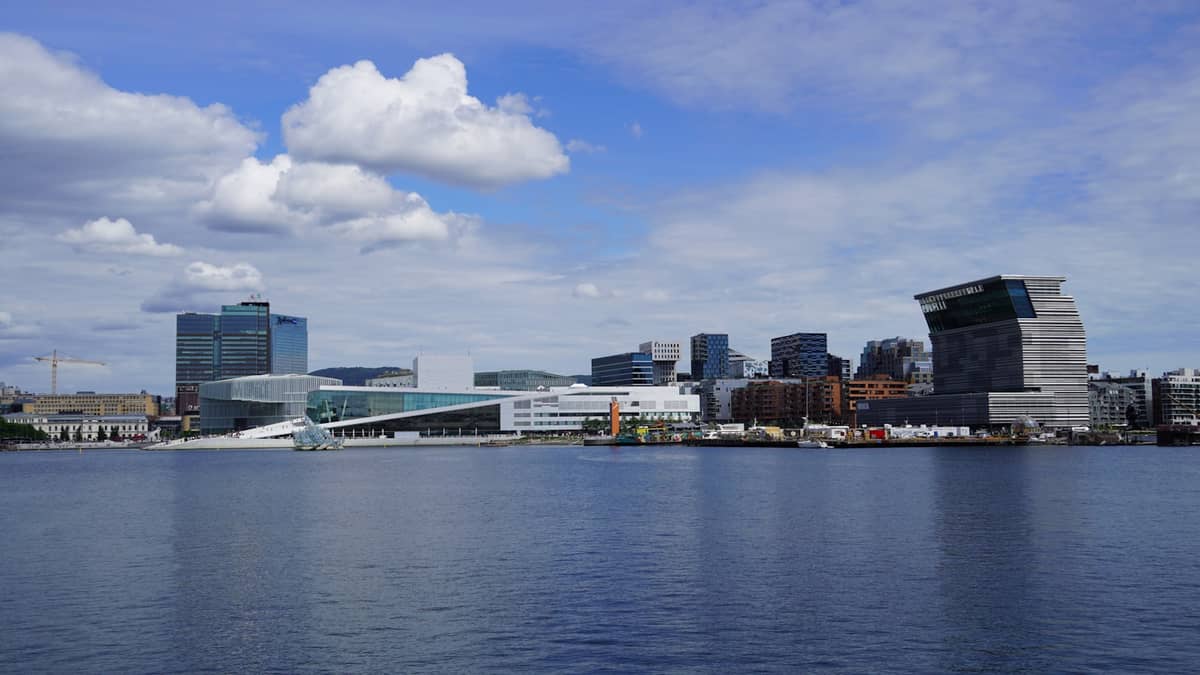 Wide view of Oslo waterfront with Oslo Opera House and MUNCH museum, modern skyline, and harbor under a partly cloudy sky.