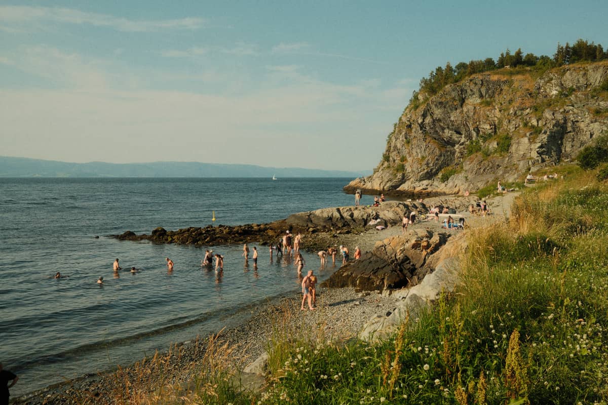 เด็กและครอบครัวว่ายน้ำและเล่นบนชายหาดหินริม Oslofjord ใต้แสงแดดฤดูร้อน พร้อมหน้าผาที่เขียวชอุ่มและน้ำสงบนิ่งเป็นฉากหลัง